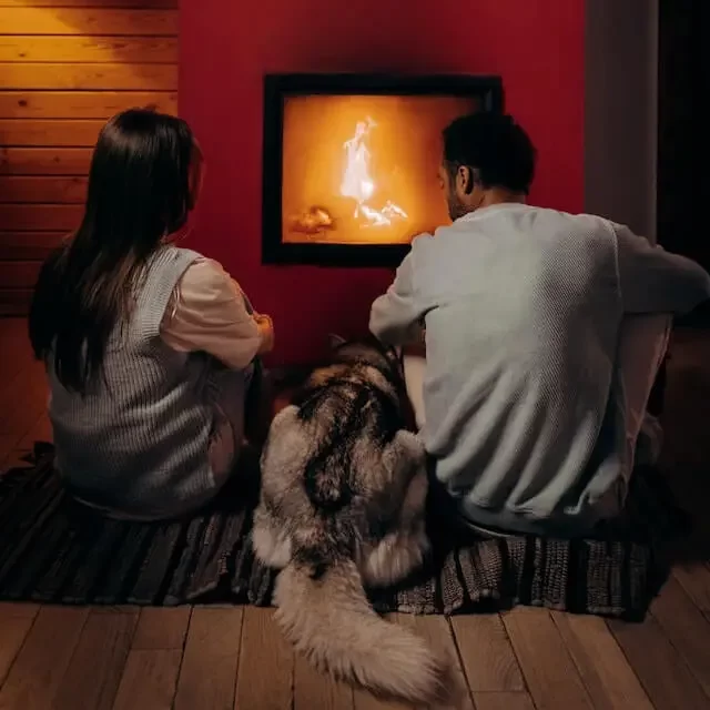 A husky sitting in between his owners in front of a cozy fireplace